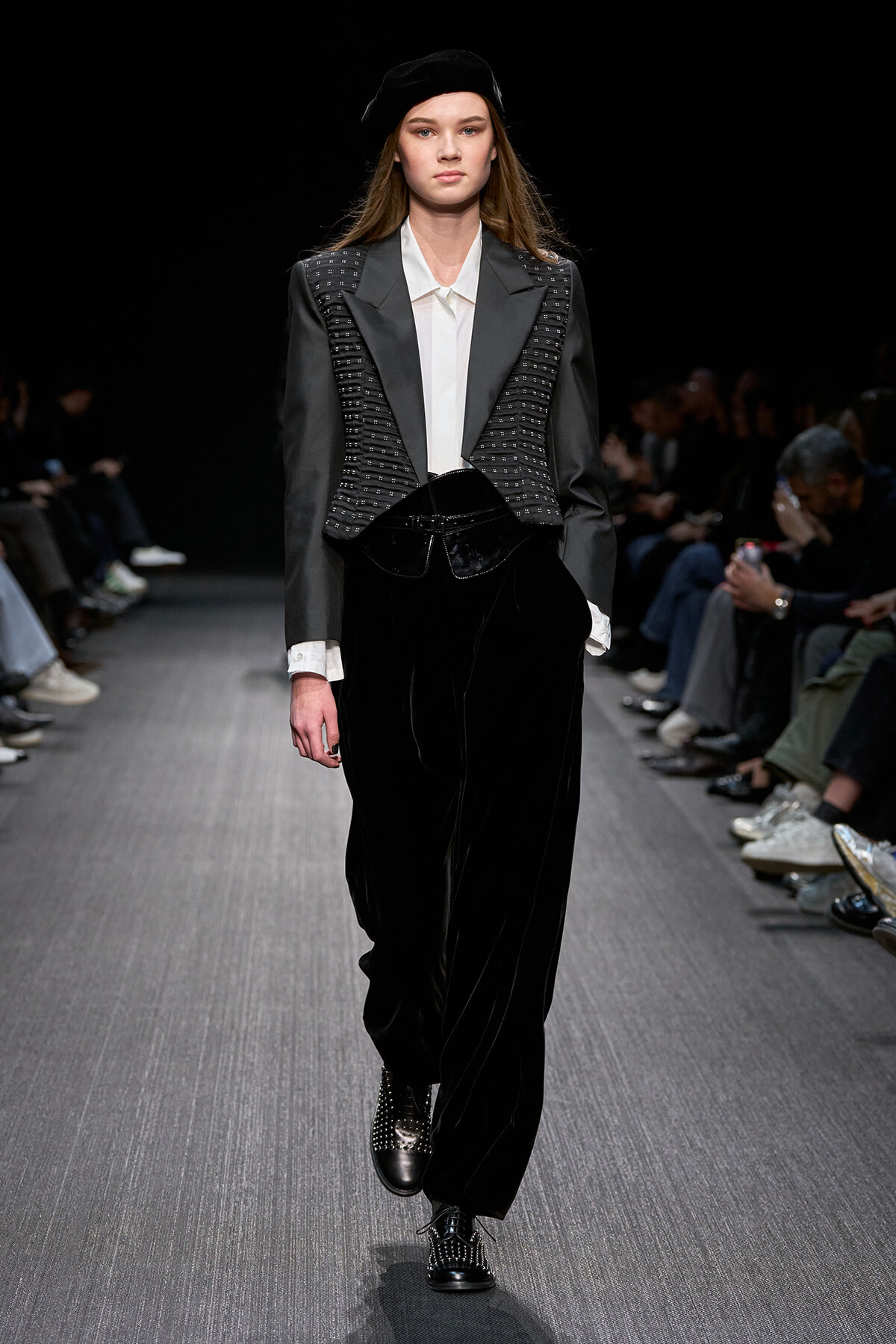 Model walking a fashion show in a gray studded blazer, white shirt, black velvet pants, and a beret on a dark runway.