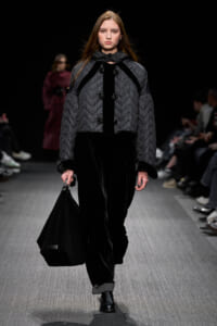 Runway model wearing a gray quilted coat with black trim and large buttons, carrying a black textured handbag down the catwalk.