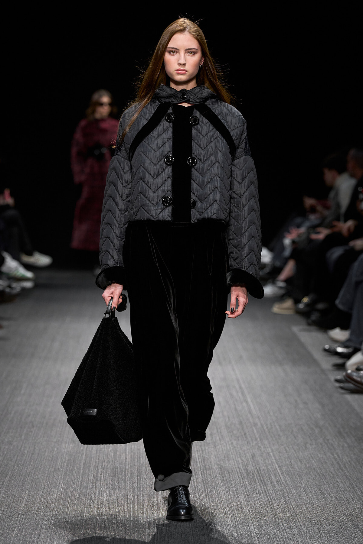 Runway model wearing a gray quilted coat with black trim and large buttons, carrying a black textured handbag down the catwalk.