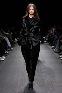 Model walks a fashion runway in a black velvet ensemble with an oversized belted jacket and matching pants, audience visible on both sides.