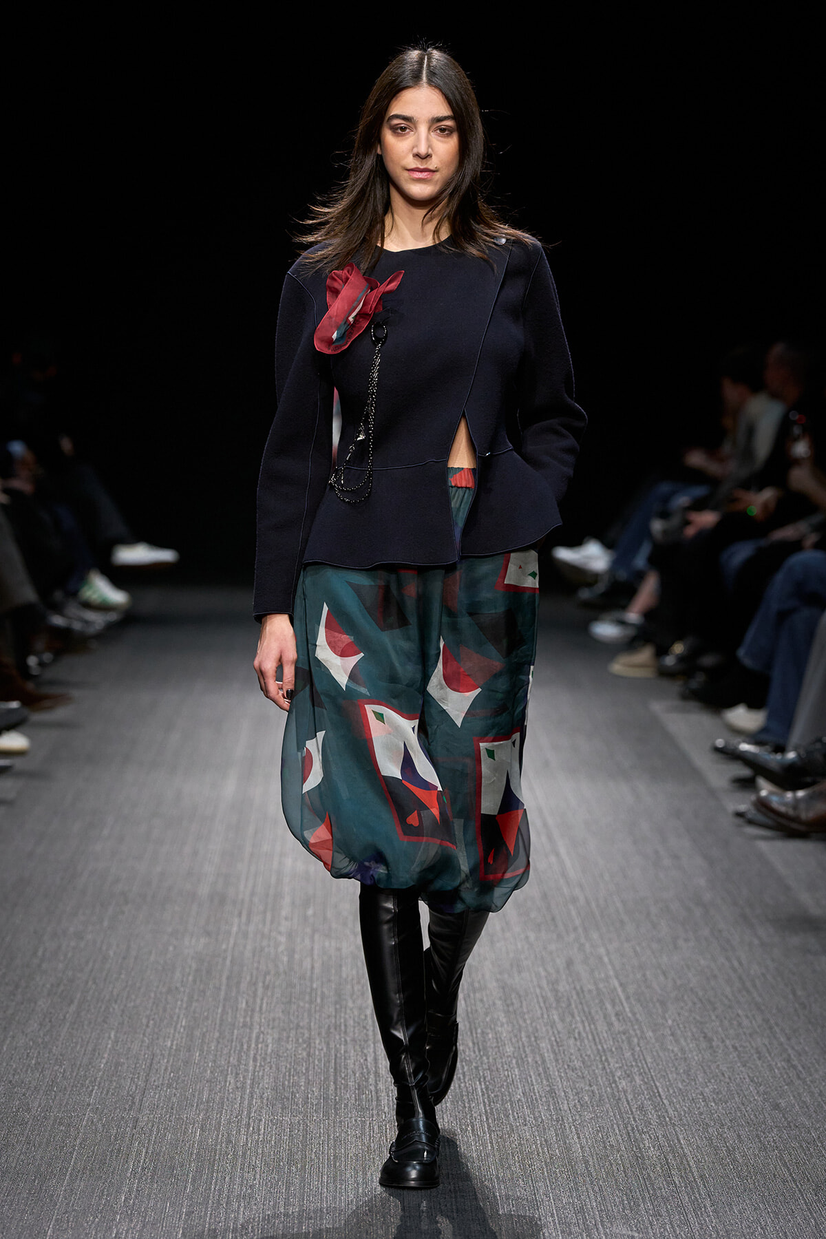 Model on a runway wearing a navy peplum-style top with a red fabric detail and a teal geometric bubble skirt, black knee-high boots.