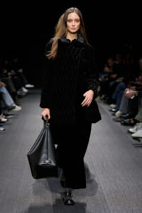 Model walking a runway in a black quilted velvet coat, carrying a large black tote on a dark catwalk.