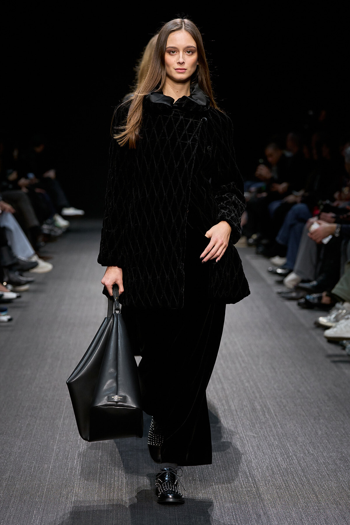 Model walking a runway in a black quilted velvet coat, carrying a large black tote on a dark catwalk.