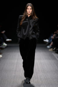 Model walks the runway in a black abstract-patterned jacket and velvet pants at a fashion show, serious expression.