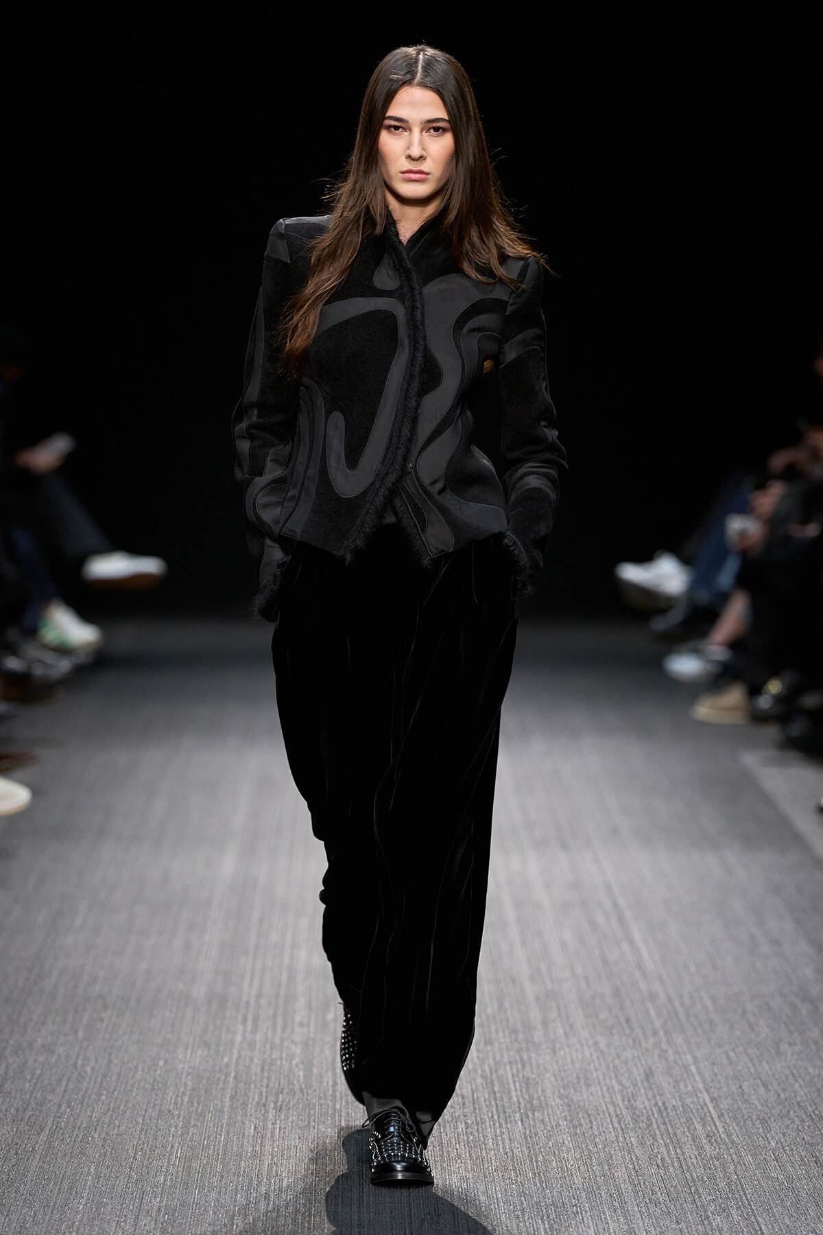 Model walks the runway in a black abstract-patterned jacket and velvet pants at a fashion show, serious expression.
