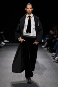 Female model walks a fashion runway in a black cape-style coat over a white shirt, black tie, and black velvet pants.