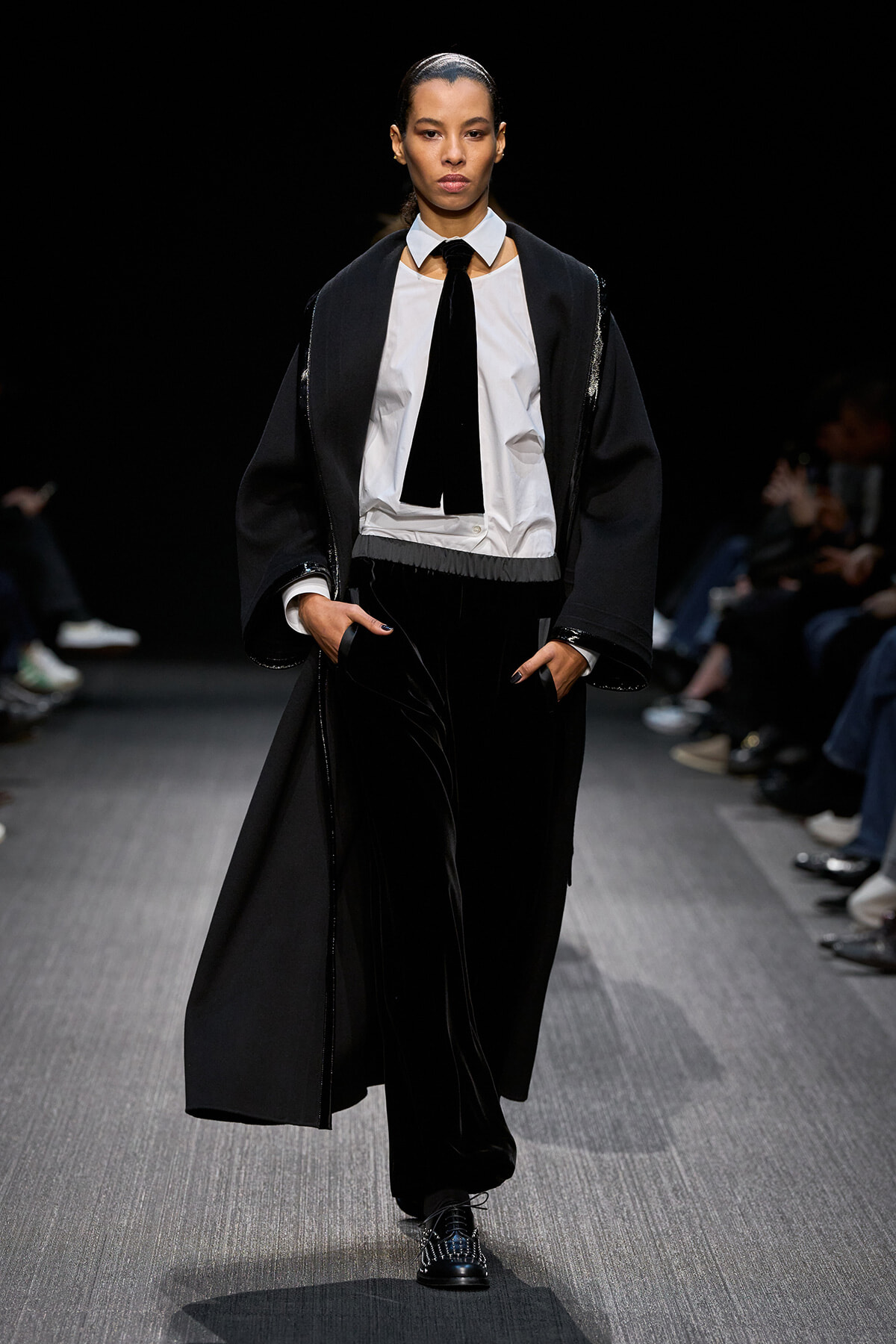 Female model walks a fashion runway in a black cape-style coat over a white shirt, black tie, and black velvet pants.