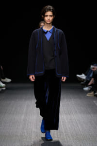 Model walking a runway in a navy cardigan with blue piping over a blue collared top and black inner layer, matching navy pants and bright blue shoes.