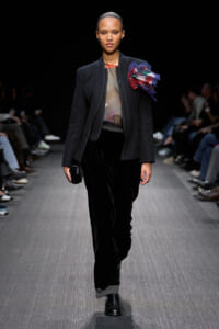 Model on a runway wearing a dark blazer over a sheer top with a large colorful bow on the shoulder, black velvet pants, and black lace-up boots.