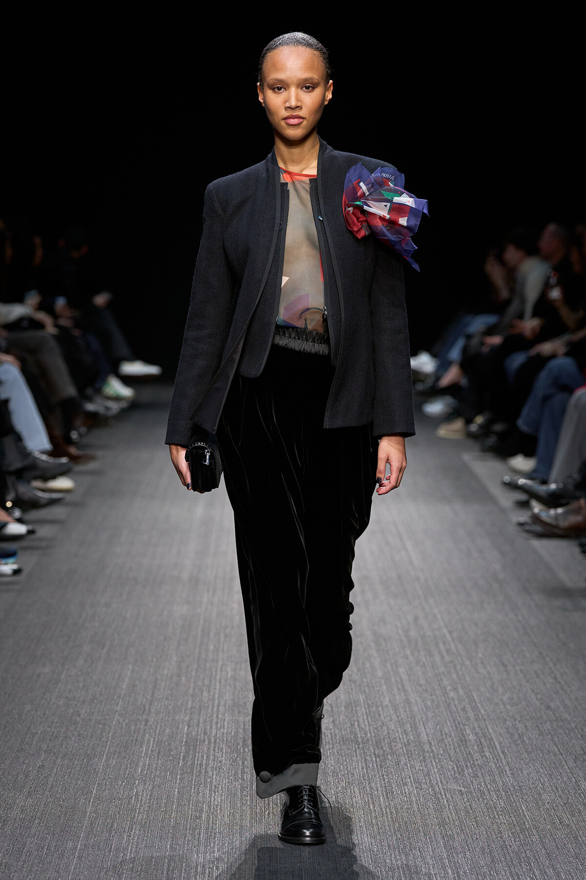 Model on a runway wearing a dark blazer over a sheer top with a large colorful bow on the shoulder, black velvet pants, and black lace-up boots.