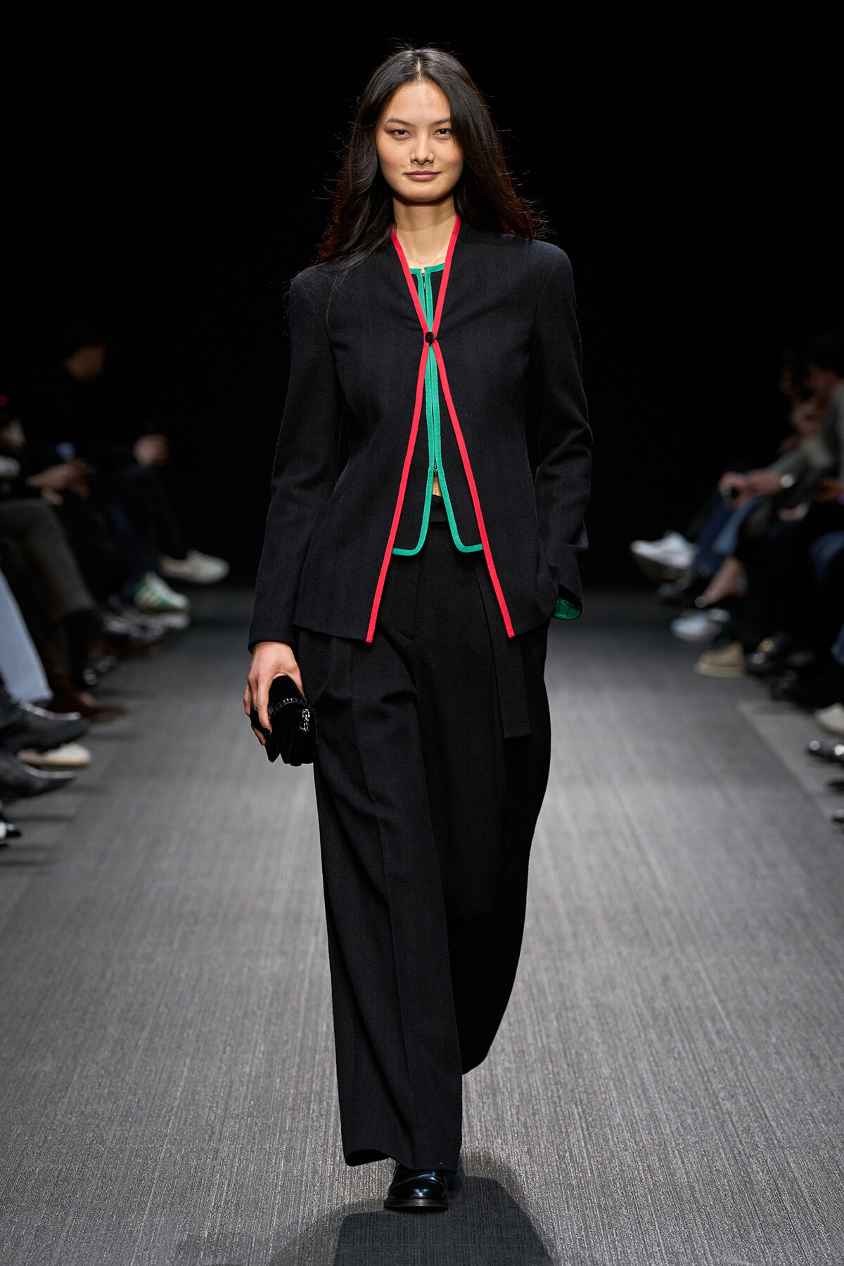 Model walks a runway wearing a black tailored blazer with bright pink piping and green accents, paired with wide-leg black pants and a small clutch.