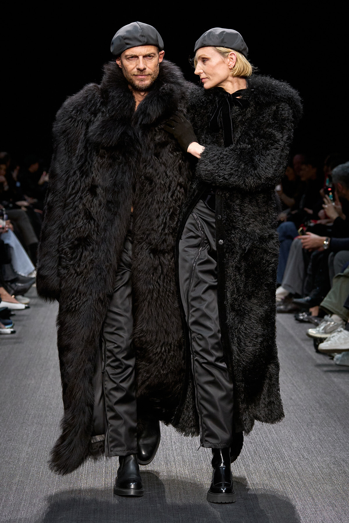 Two models walk a runway in matching black fur coats and gray berets, close together as they pose for the crowd.