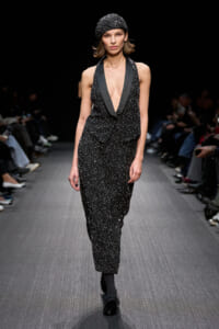Model on a runway wearing a black sequined outfit with a deep V-neck and matching cap.