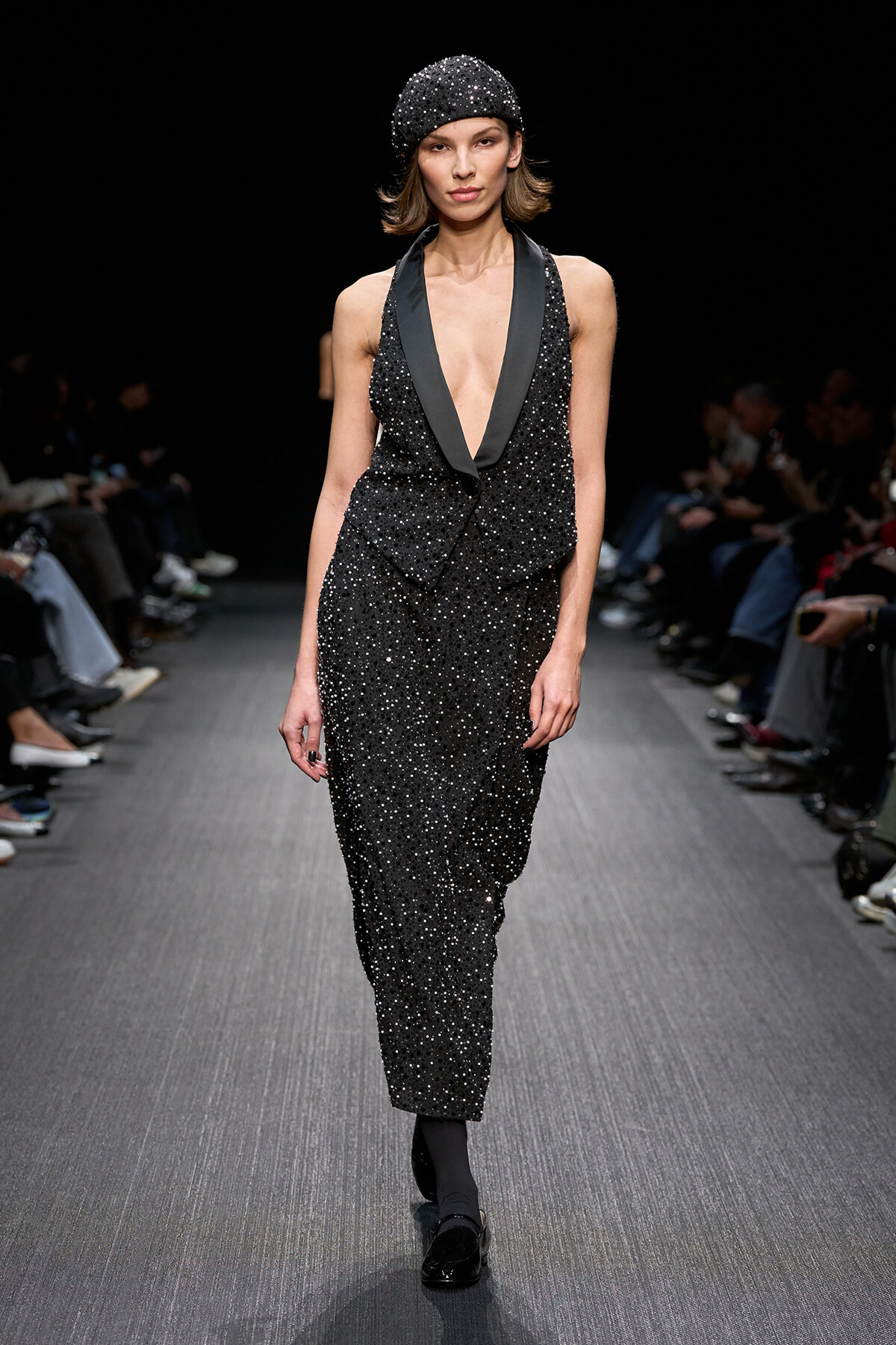 Model on a runway wearing a black sequined outfit with a deep V-neck and matching cap.