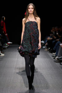 Model walks a fashion show wearing a strapless black bubble dress with multicolor curved lines in red, green, and blue, paired with black tights and heeled boots.