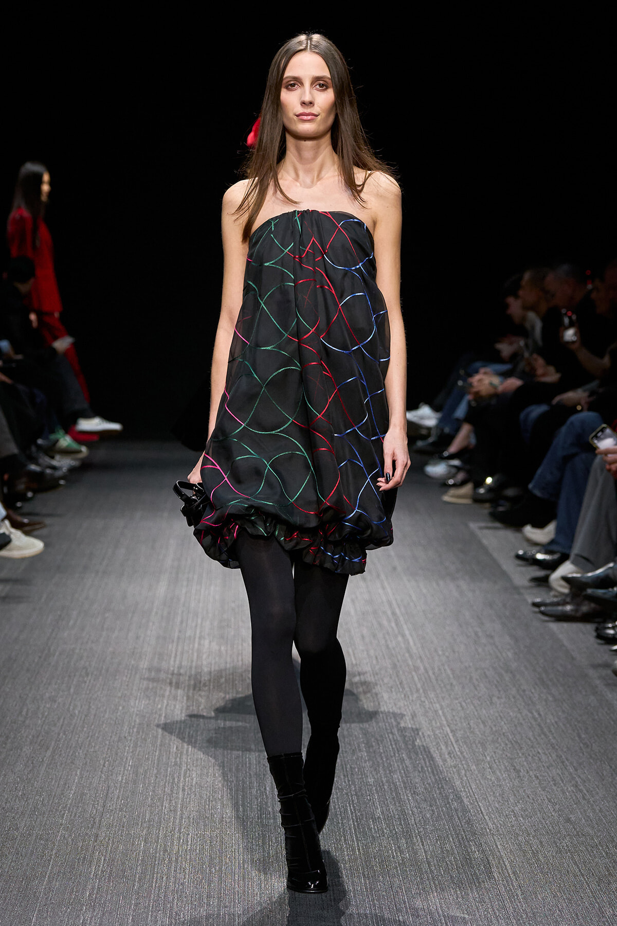 Model walks a fashion show wearing a strapless black bubble dress with multicolor curved lines in red, green, and blue, paired with black tights and heeled boots.