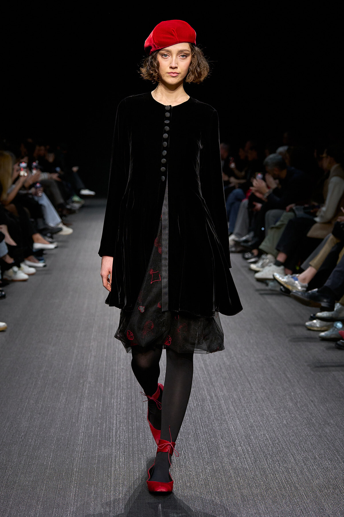 Model walks a runway in a black velvet coat with a red beret and red lace-up shoes, audience in the background.