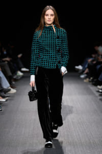 Model walking a fashion show runway in a teal grid-pattern jacket with white cuffs and black velvet wide-leg pants, carrying a small black handbag.