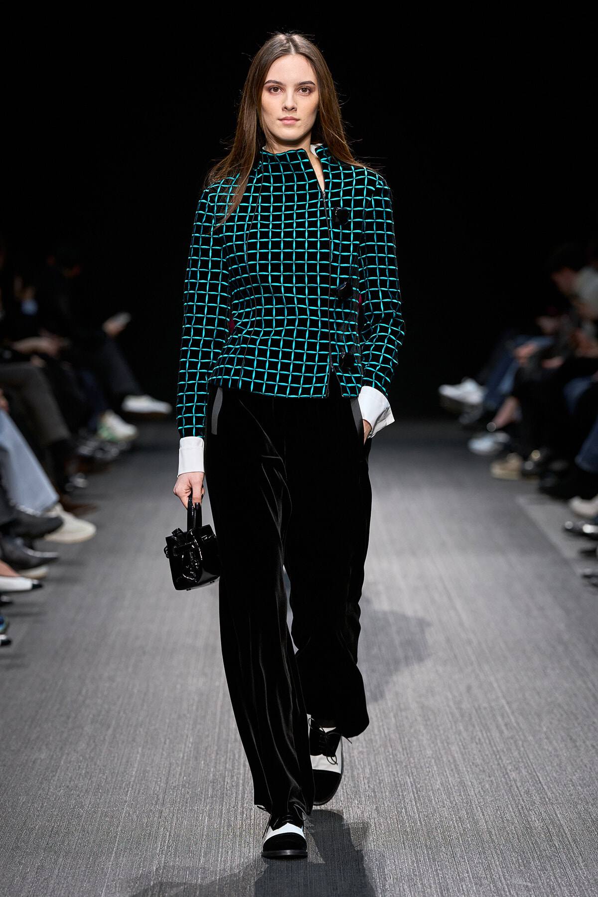 Model walking a fashion show runway in a teal grid-pattern jacket with white cuffs and black velvet wide-leg pants, carrying a small black handbag.
