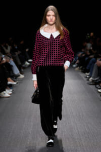Model walks the runway in a black velvet wide-leg pant suit with a pink grid-pattern top and a white Peter Pan collar; small black handbag in hand.