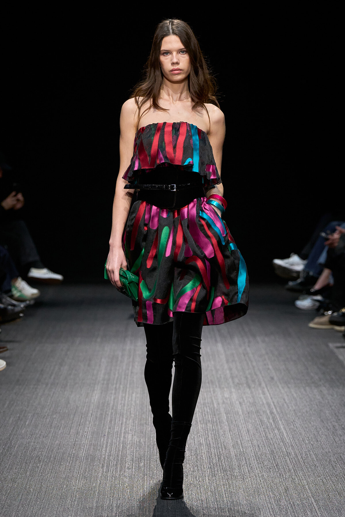 Model walks the runway in a strapless, multicolored striped dress with a black belt, black over-the-knee boots, and a green clutch.