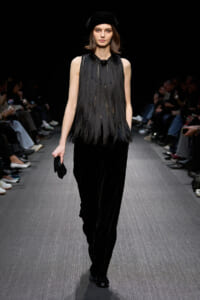 Model walks the runway in a black fringe sleeveless top with velvet wide-leg pants, accessorized by a black hat and small clutch on the side
