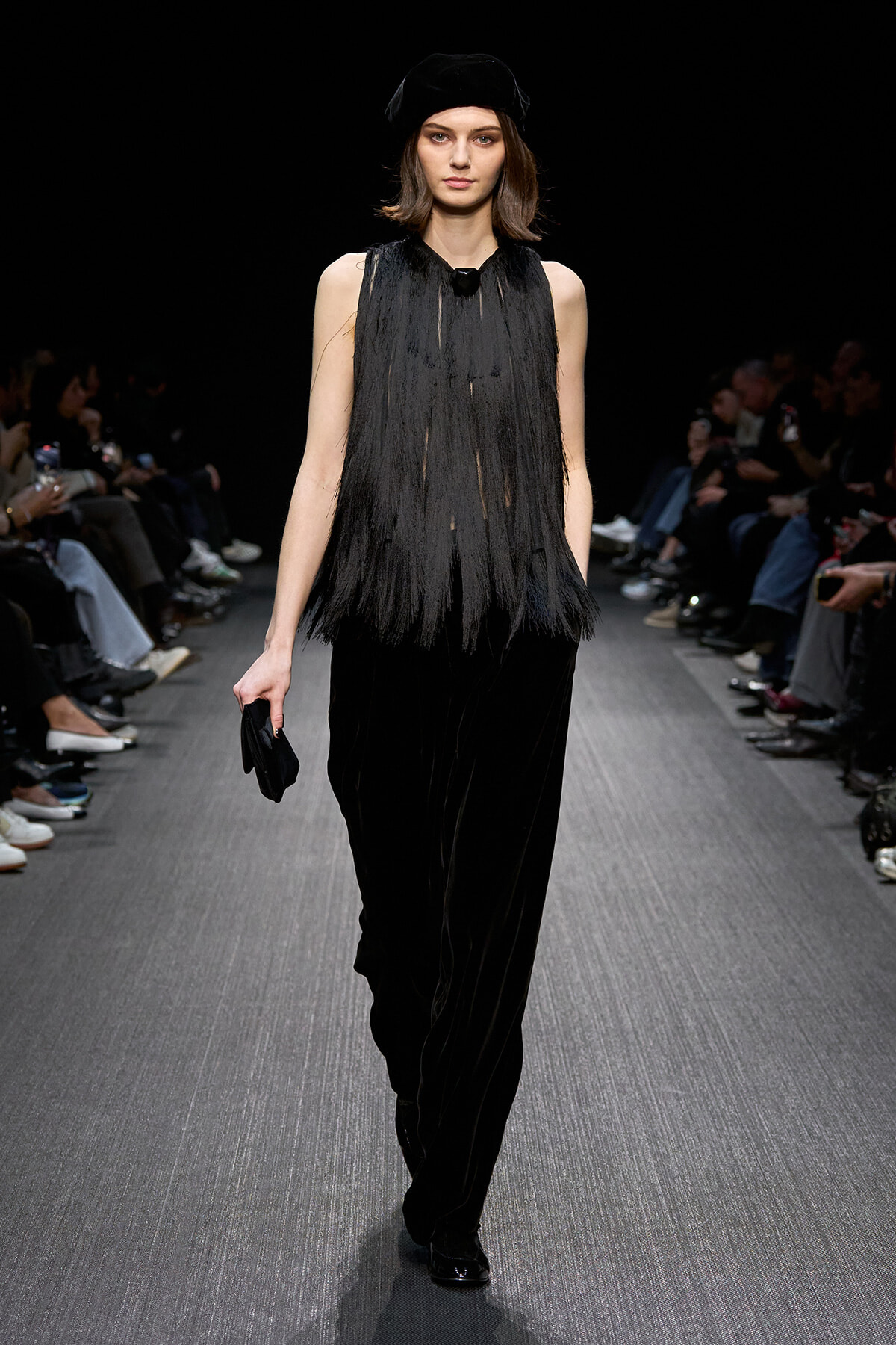 Model walks the runway in a black fringe sleeveless top with velvet wide-leg pants, accessorized by a black hat and small clutch on the side