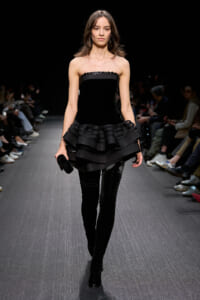 Model walking a runway in a black strapless dress with a layered ruffle peplum, black thigh-high boots, and a small black clutch.