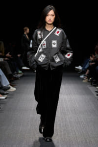 Model walking the runway in a black bomber jacket covered with sewn-on playing-card patches, paired with black wide-leg pants and studded shoes, under a dark backdrop.