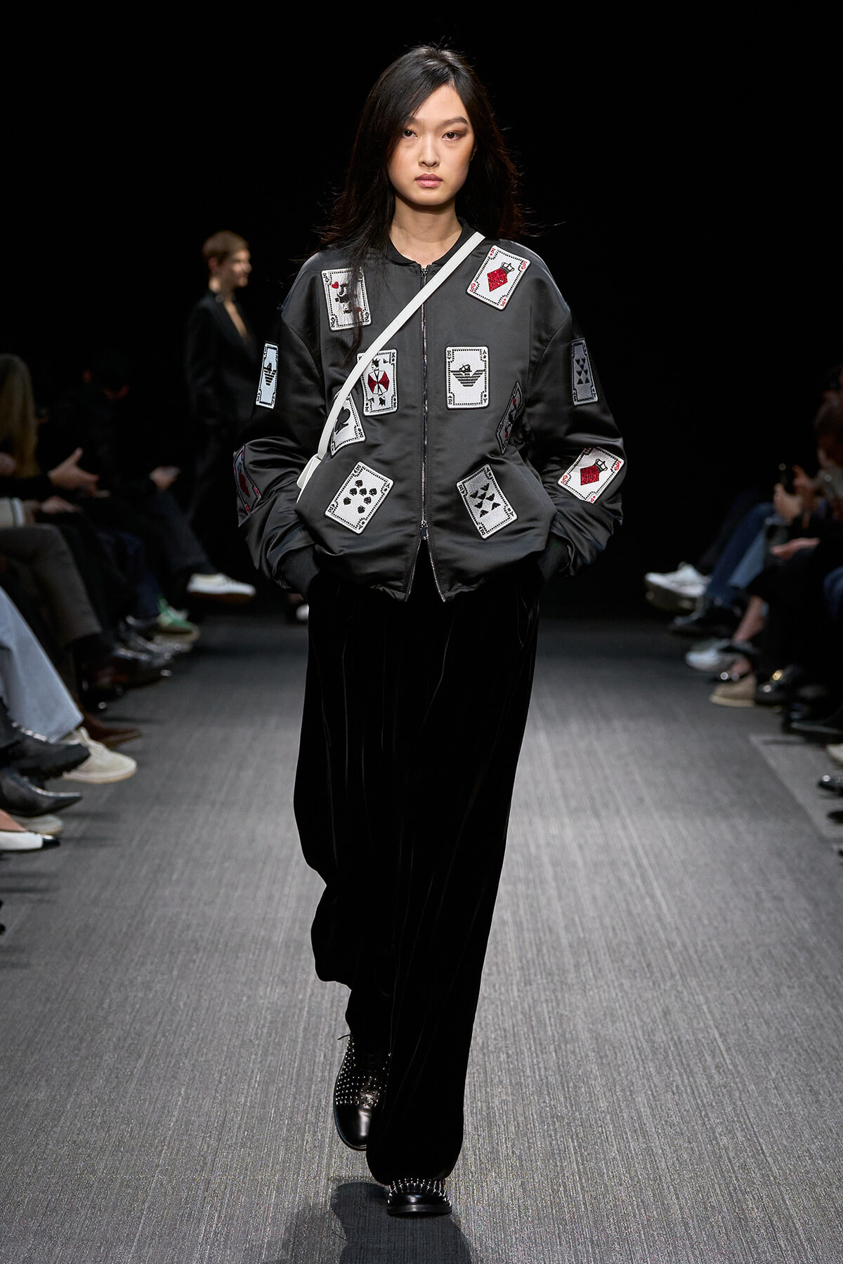 Model walking the runway in a black bomber jacket covered with sewn-on playing-card patches, paired with black wide-leg pants and studded shoes, under a dark backdrop.