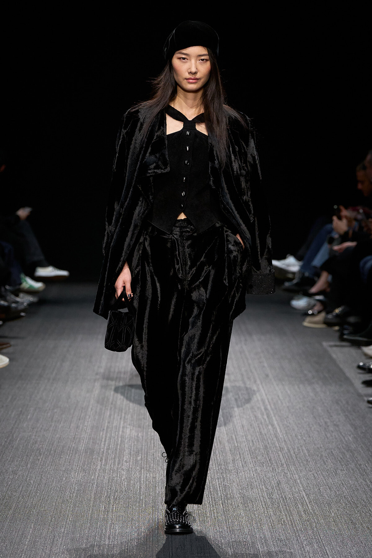 Model walks a runway in an all-black velvet ensemble: long coat, vest, loose pants, and a black hat, carrying a small black bag.