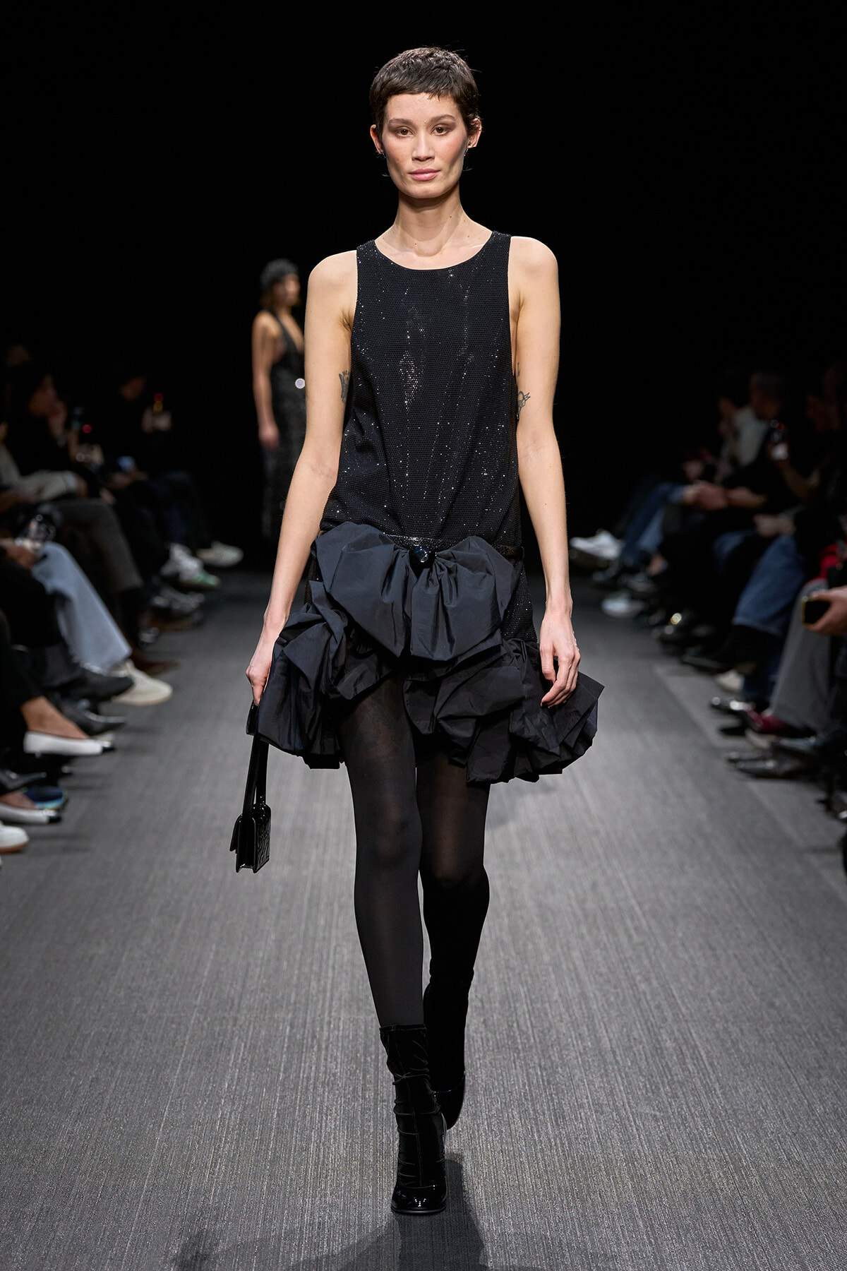Model walking a runway in a sleeveless black sequin top and a voluminous black ruffled skirt, carrying a small black handbag.