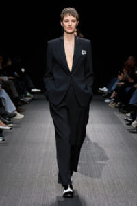 Female model wearing a dark, oversized black pantsuit with a plunging blazer on a runway, hands in pockets, silver earrings visible.