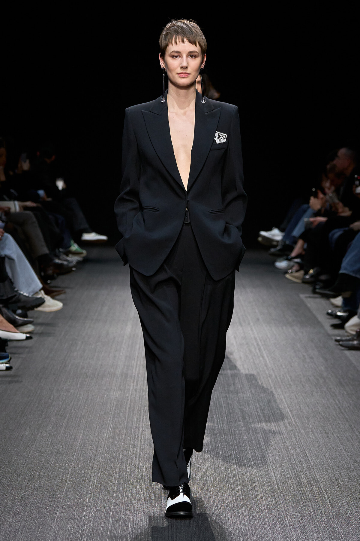 Female model wearing a dark, oversized black pantsuit with a plunging blazer on a runway, hands in pockets, silver earrings visible.
