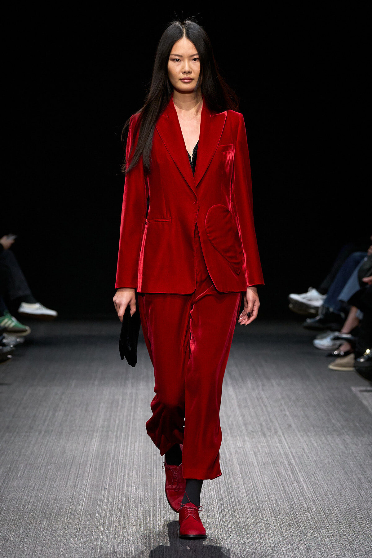 Model walking a fashion runway in a red velvet suit: tailored blazer with a curved pocket and matching trousers, black top underneath, carrying a small black clutch.
