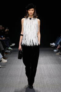 Model walks the runway in a white fringe sleeveless top, black wide-leg pants, a black beret, and a small black handbag.