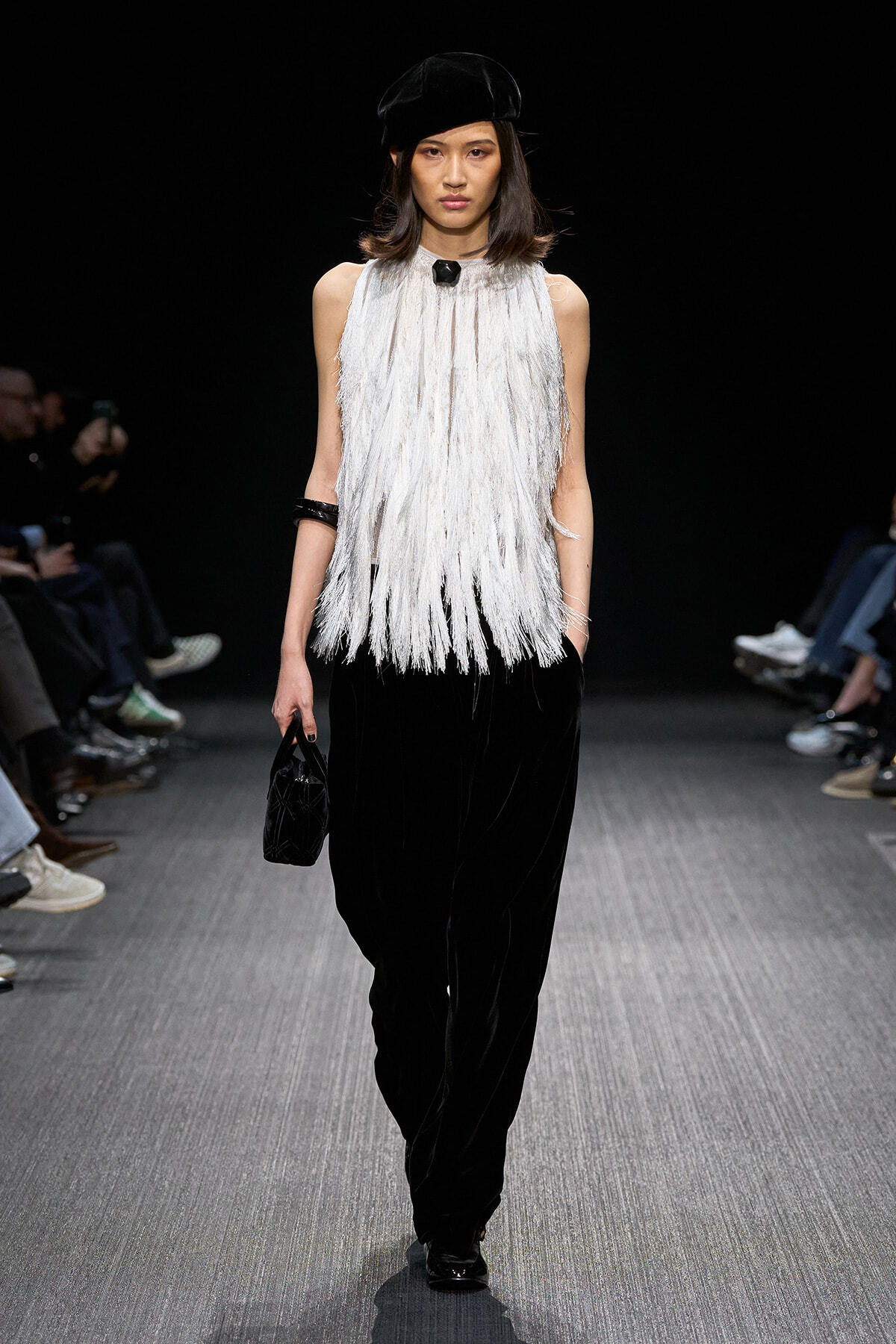 Model walks the runway in a white fringe sleeveless top, black wide-leg pants, a black beret, and a small black handbag.