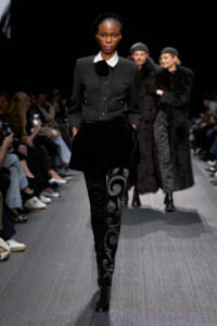 Model walks the runway in a black tailored jacket with a white collar, velvet A-line skirt, and swirling-patterned tights, audience shown on sides.