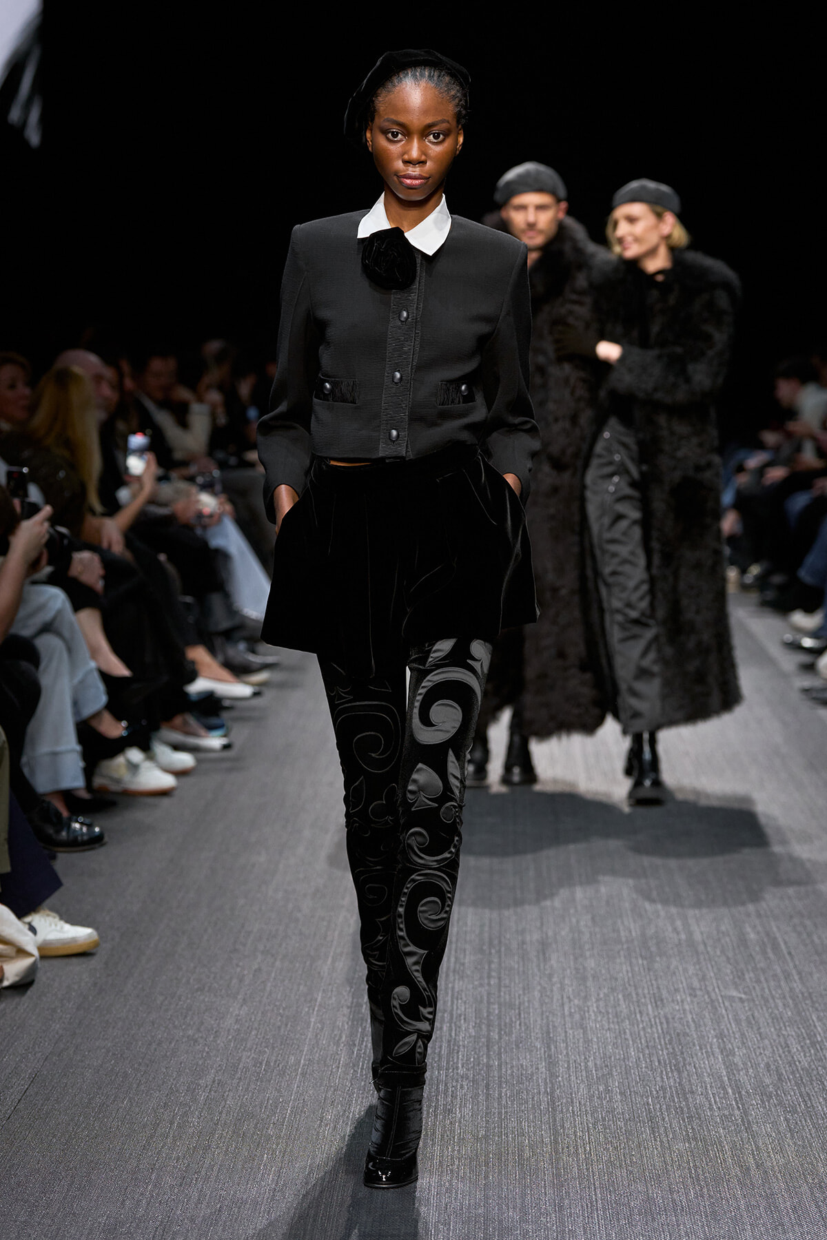 Model walks the runway in a black tailored jacket with a white collar, velvet A-line skirt, and swirling-patterned tights, audience shown on sides.
