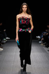 Model walks runway in strapless multicolor ruffled top with black velvet pants and belt, holding a blue clutch.