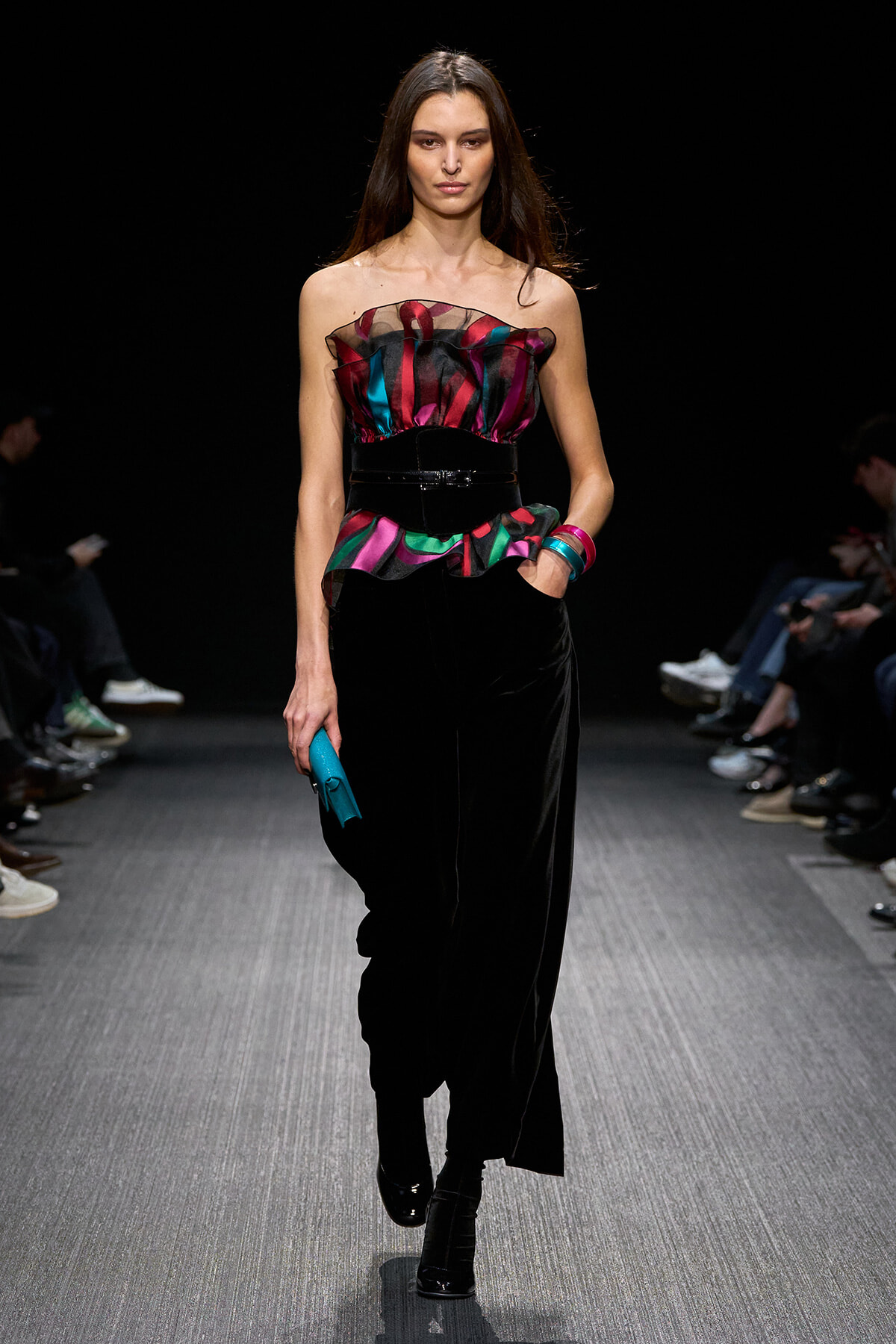 Model walks runway in strapless multicolor ruffled top with black velvet pants and belt, holding a blue clutch.