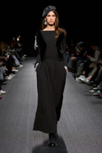 Model walks a black-clad runway look: beret, long-sleeve top, velvet-trim vest, wide-leg pants, and studded shoes.