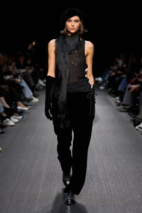 Model walks a runway in all-black ensemble: sequined sleeveless top, wide-leg velvet pants, long black scarf, gloves and a black beret.