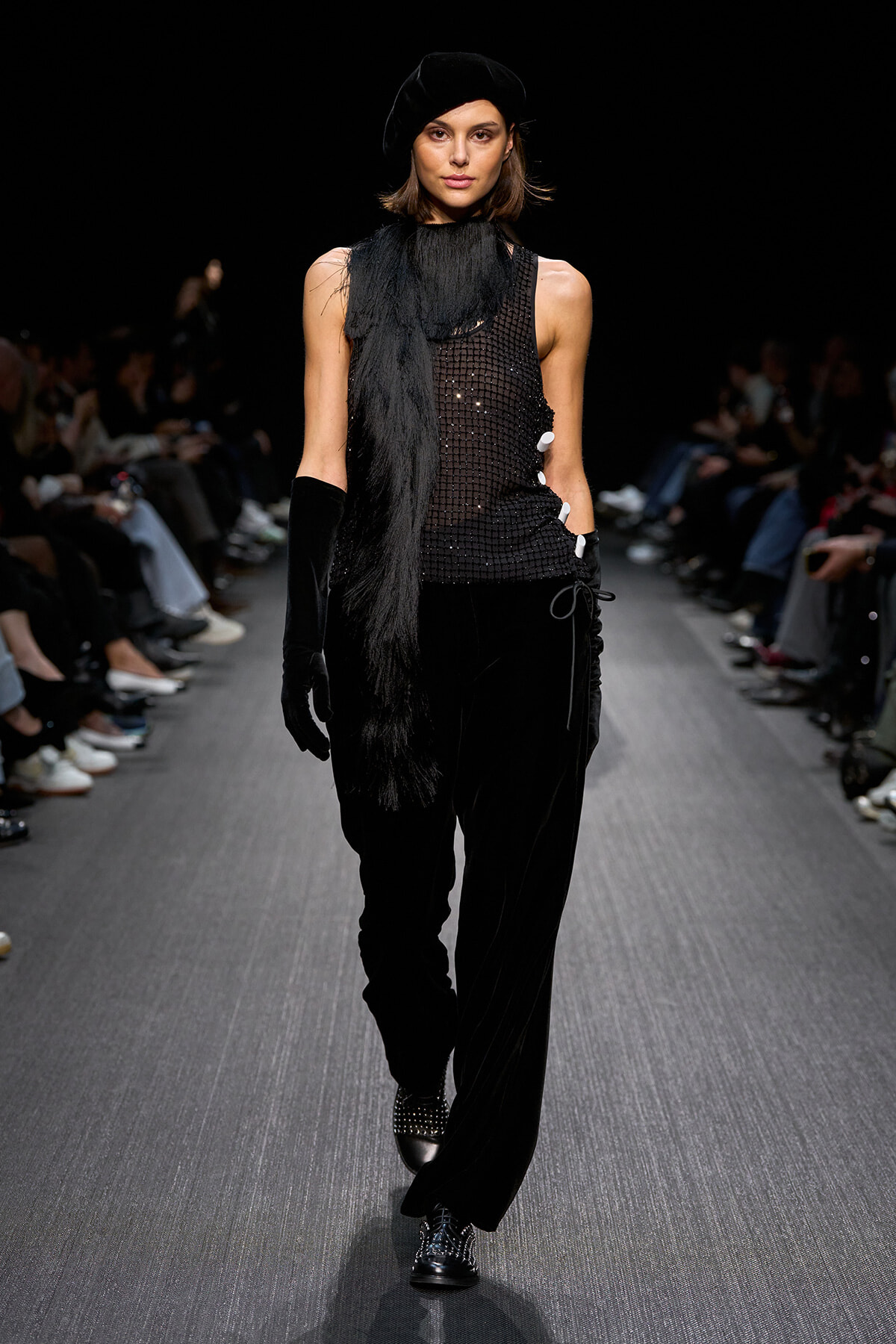 Model walks a runway in all-black ensemble: sequined sleeveless top, wide-leg velvet pants, long black scarf, gloves and a black beret.