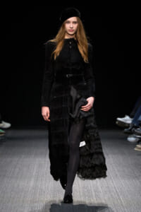 Female model walks the runway in a black velvet ensemble featuring a long textured coat, wide belt, and beret.