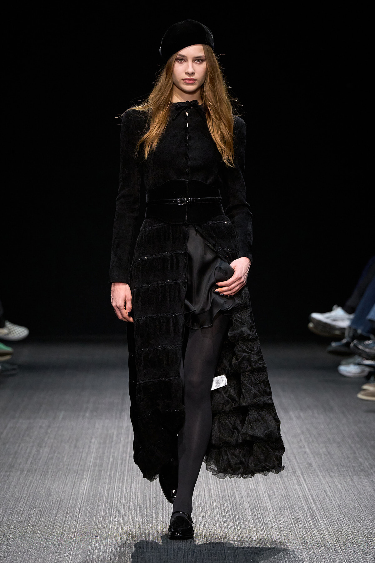 Female model walks the runway in a black velvet ensemble featuring a long textured coat, wide belt, and beret.