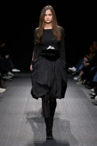 Female model walks a fashion runway in a black long-sleeve top and a voluminous, knee-length black skirt with a wide belt and studded boots.