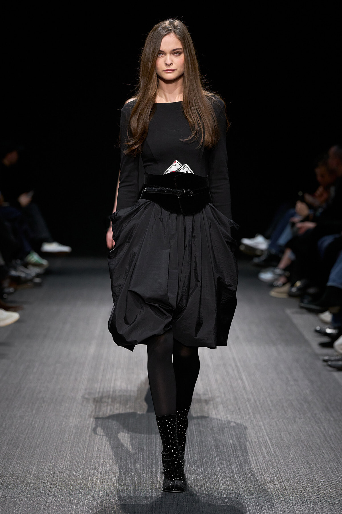 Female model walks a fashion runway in a black long-sleeve top and a voluminous, knee-length black skirt with a wide belt and studded boots.