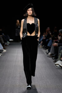 Model on a runway wearing a black velvet heart-shaped crop top, satin vest, wide-leg pants, and a black beret.