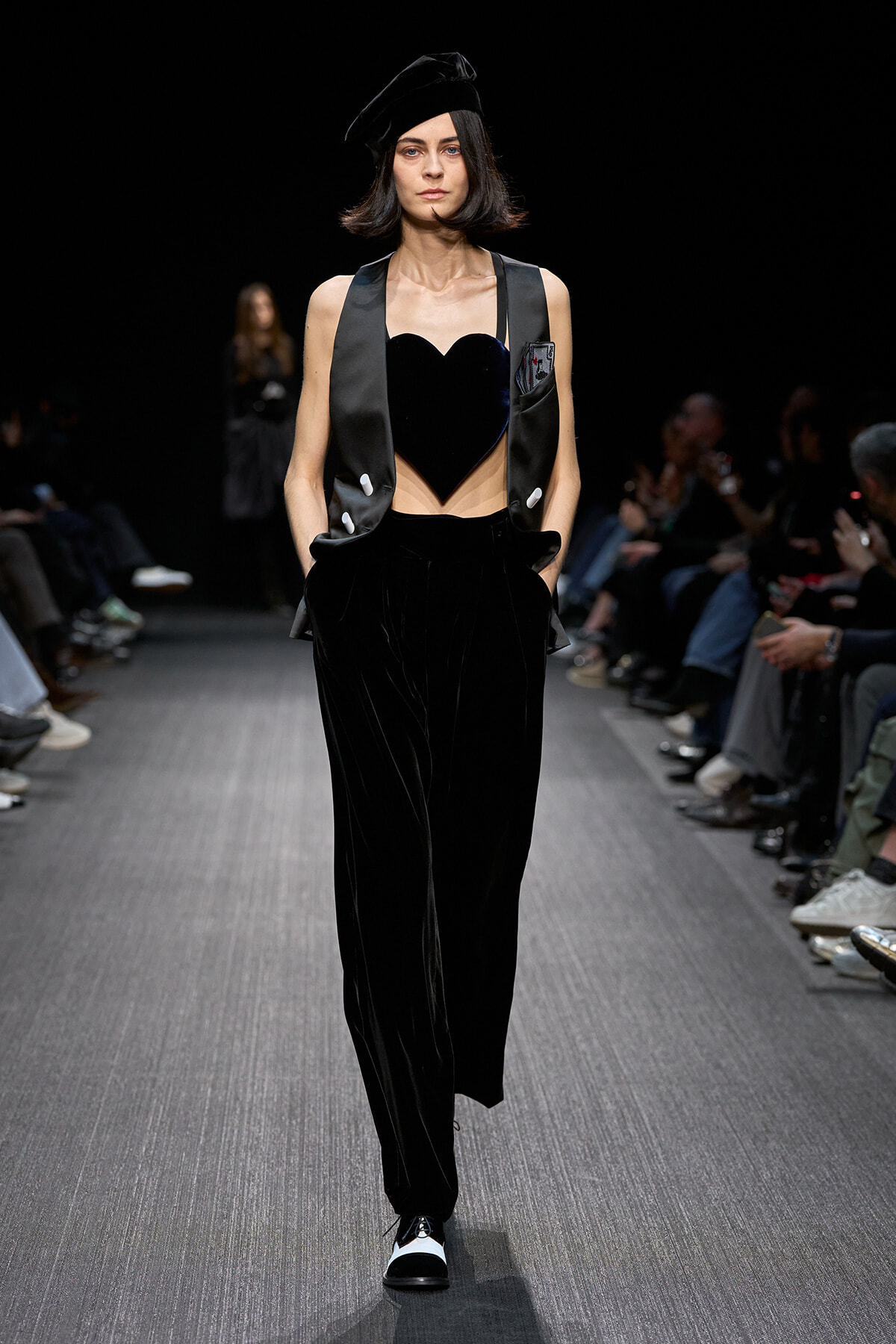 Model on a runway wearing a black velvet heart-shaped crop top, satin vest, wide-leg pants, and a black beret.